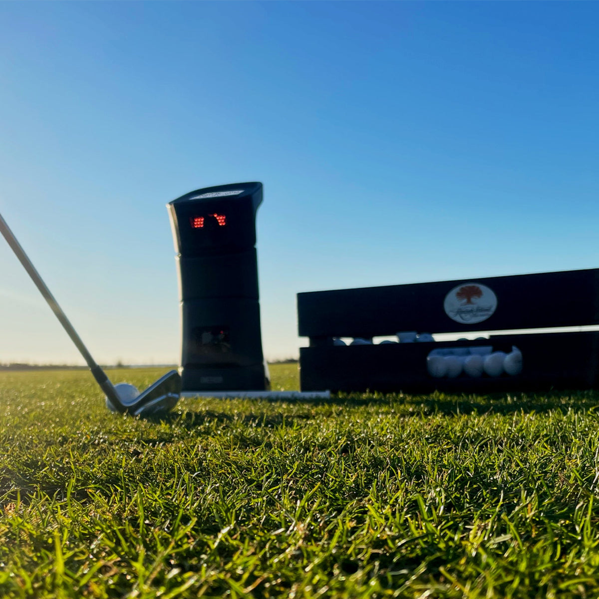 Uneekor Eye Mini launch monitor placed on outdoor grass, with a golf club and golf balls nearby