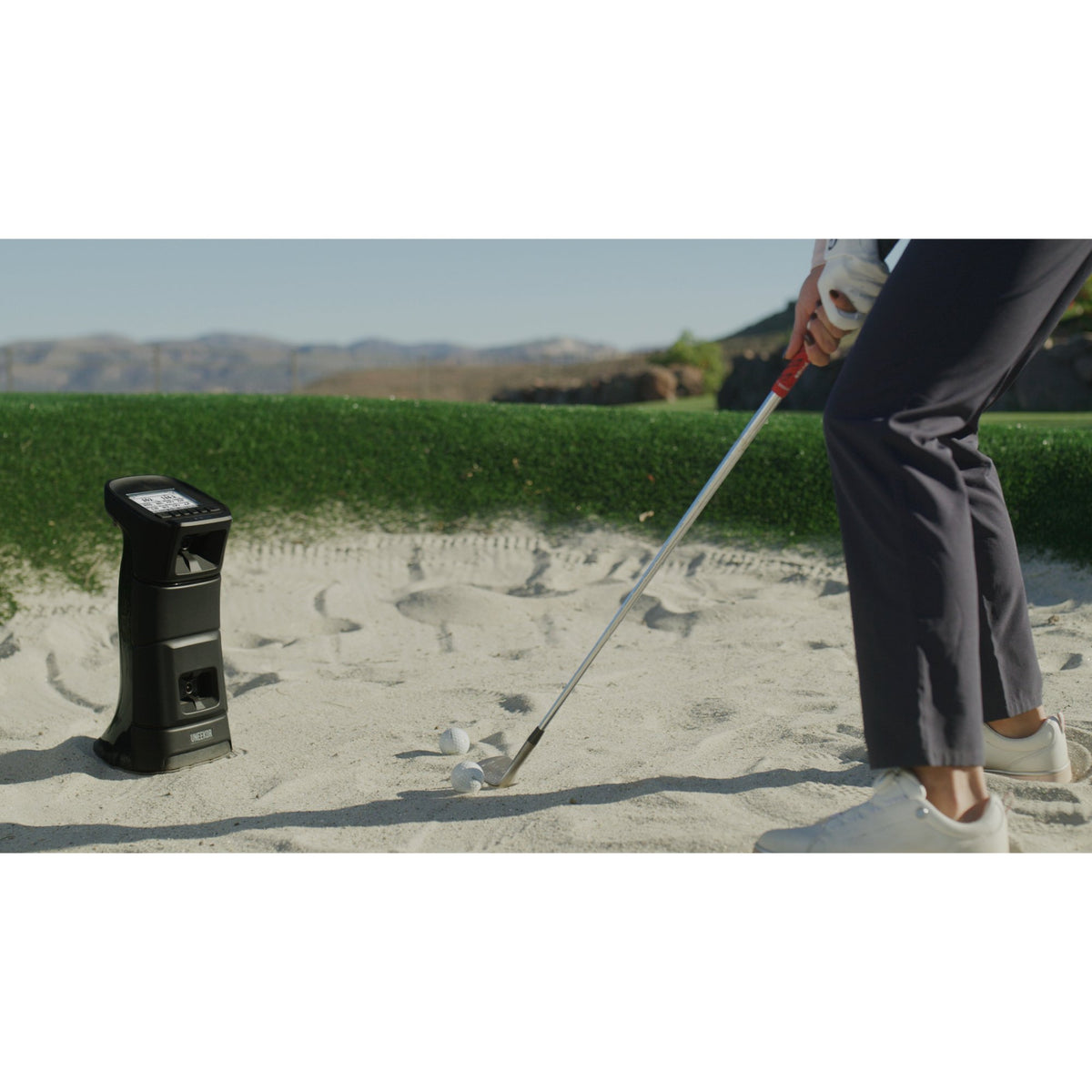 A golfer hitting a ball from a sand bunker with the Uneekor Eye Mini launch monitor nearby.